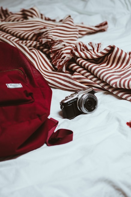 A red backpack and a striped maroon and white scarf rest on a white surface. A compact camera is placed beside them, suggesting themes of travel or exploration.