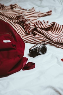 A red backpack and a striped maroon and white scarf rest on a white surface. A compact camera is placed beside them, suggesting themes of travel or exploration.