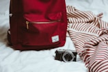 A red backpack is placed on a white bed next to a camera and a striped red and white blanket. The camera is positioned in front of the backpack, while the blanket is casually draped.