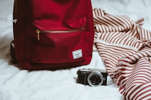 A red backpack is placed on a white bed next to a camera and a striped red and white blanket. The camera is positioned in front of the backpack, while the blanket is casually draped.