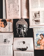 A collection of open books and a bust of a Buddha positioned centrally, creating a symmetrical and balanced composition. The books feature Bengali text and illustrations, suggesting historical or literary content. The scene is rich in cultural and spiritual elements.