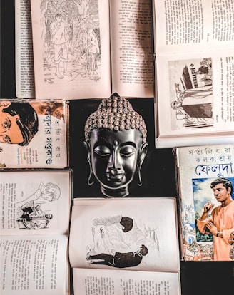 A collection of open books and a bust of a Buddha positioned centrally, creating a symmetrical and balanced composition. The books feature Bengali text and illustrations, suggesting historical or literary content. The scene is rich in cultural and spiritual elements.