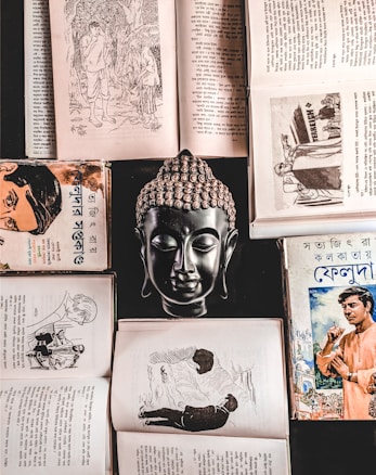 A collection of open books and a bust of a Buddha positioned centrally, creating a symmetrical and balanced composition. The books feature Bengali text and illustrations, suggesting historical or literary content. The scene is rich in cultural and spiritual elements.