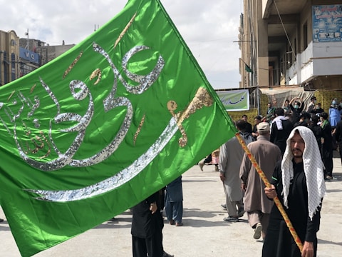 A man with a headscarf holds a large green flag with Arabic script and a sword design. The background shows a crowd of people walking on a city street with buildings lining the sides. The atmosphere suggests a public procession or gathering.