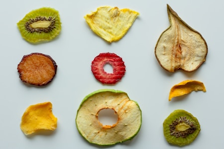 An assortment of dried fruit slices, including kiwi, pineapple, pear, apple, strawberry, and mango, is arranged on a light background in a grid-like pattern. The fruit slices vary in color, texture, and size.