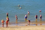 Several people wearing swim caps and swimsuits are in the water near a sandy beach. Some are wading while others are adjusting their caps or preparing to swim.