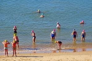 Several people wearing swim caps and swimsuits are in the water near a sandy beach. Some are wading while others are adjusting their caps or preparing to swim.