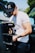 A man wearing a black cap and a white t-shirt operates a professional video camera rig. He appears focused on his equipment, which has additional monitors and hardware attached. The background shows car doors open and some greenery.
