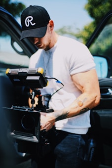 A man wearing a black cap and a white t-shirt operates a professional video camera rig. He appears focused on his equipment, which has additional monitors and hardware attached. The background shows car doors open and some greenery.