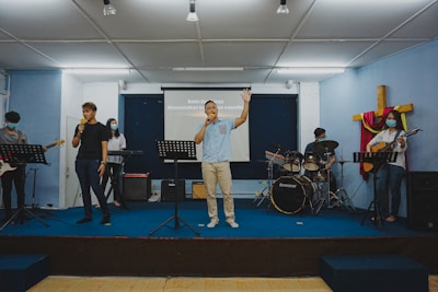 Several people are performing music on a stage, each with different instruments. Two individuals are holding microphones, one is playing the keyboard, another person is on the drums, and another is playing a guitar. A cross decorated with fabric is visible in the background, suggesting a religious setting. They appear to be in a room with a projector screen displaying text.