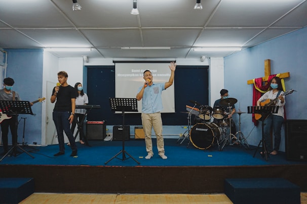 Several people are performing music on a stage, each with different instruments. Two individuals are holding microphones, one is playing the keyboard, another person is on the drums, and another is playing a guitar. A cross decorated with fabric is visible in the background, suggesting a religious setting. They appear to be in a room with a projector screen displaying text.