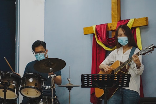 A man and a woman, both wearing face masks, are playing musical instruments in a room with a large wooden cross draped in red and yellow cloth on the wall. The man is playing drums, while the woman is playing an acoustic guitar and standing in front of a music stand.