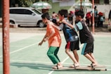 Three boys are playing a team-building or coordination game on a green outdoor sports court. They are balancing on wooden planks with their arms on each other's shoulders. There are other people and vehicles in the background.