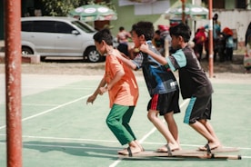 Three boys are playing a team-building or coordination game on a green outdoor sports court. They are balancing on wooden planks with their arms on each other's shoulders. There are other people and vehicles in the background.