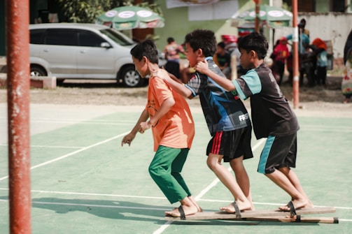 Three boys are playing a team-building or coordination game on a green outdoor sports court. They are balancing on wooden planks with their arms on each other's shoulders. There are other people and vehicles in the background.