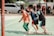 Three boys are playing a team-building or coordination game on a green outdoor sports court. They are balancing on wooden planks with their arms on each other's shoulders. There are other people and vehicles in the background.