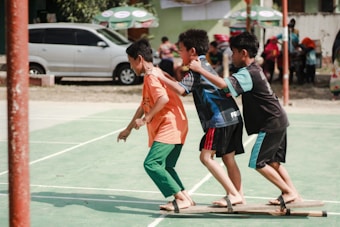 Three boys are playing a team-building or coordination game on a green outdoor sports court. They are balancing on wooden planks with their arms on each other's shoulders. There are other people and vehicles in the background.