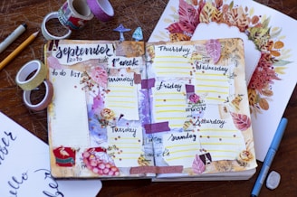 A decorated open journal features hand-drawn weekly planning pages for September 2020. Surrounding the journal are various stationery items, including rolls of washi tape, pencils, pens, and a drawing of fall leaves. The journal pages are adorned with floral and leaf decorations, sticky notes with days of the week, and glittery tape.