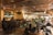A warmly lit interior of a bar or restaurant featuring a live band performing on one side. The audience, seated at tables and bar stools, enjoys the music. The space is decorated with intricate ceiling designs and a mounted animal head on the wall. The atmosphere is cozy and lively with social interactions throughout.