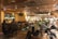 A warmly lit interior of a bar or restaurant featuring a live band performing on one side. The audience, seated at tables and bar stools, enjoys the music. The space is decorated with intricate ceiling designs and a mounted animal head on the wall. The atmosphere is cozy and lively with social interactions throughout.