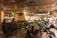 A warmly lit interior of a bar or restaurant featuring a live band performing on one side. The audience, seated at tables and bar stools, enjoys the music. The space is decorated with intricate ceiling designs and a mounted animal head on the wall. The atmosphere is cozy and lively with social interactions throughout.
