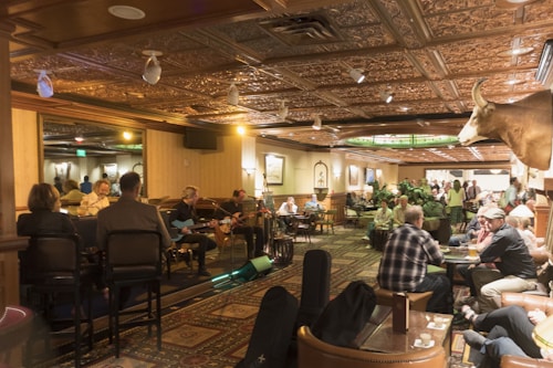 A warmly lit interior of a bar or restaurant featuring a live band performing on one side. The audience, seated at tables and bar stools, enjoys the music. The space is decorated with intricate ceiling designs and a mounted animal head on the wall. The atmosphere is cozy and lively with social interactions throughout.