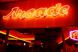 Bright neon signage displaying the word 'Arcade' in glowing red letters with a retro aesthetic. Surrounding environment shows dim lighting, with a neon 'Open' sign visible, suggesting the entrance to an entertainment venue.