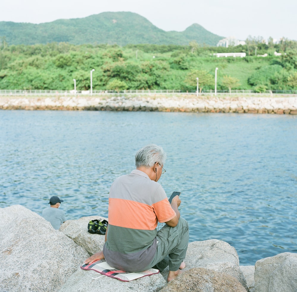 man in orange t-shirt and gray pants sitting on gray rock near body of water