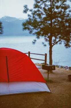A red and white tent is set up next to a body of water, with a wooden fence and a small grill nearby. A large coniferous tree stands beside the tent, and in the background, mountains are partially covered in snow.