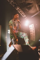 A performer is passionately singing on stage, holding a microphone. They are dressed in a colorful outfit with camouflaged pants and a printed shirt. The stage is lit with bright lights, and there are speakers and equipment visible in the background, creating a dynamic concert atmosphere.