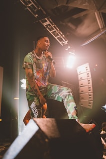 A performer is passionately singing on stage, holding a microphone. They are dressed in a colorful outfit with camouflaged pants and a printed shirt. The stage is lit with bright lights, and there are speakers and equipment visible in the background, creating a dynamic concert atmosphere.