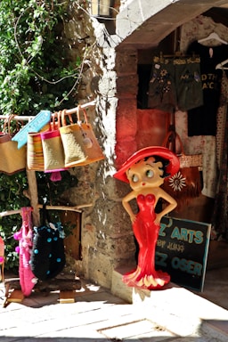 A colorful shop display features a cartoon character statue in a bright red dress and hat. Surrounding the statue are woven bags in various colors, hanging under a stone archway. The background includes olive green shorts and a black shirt with a partial design visible.