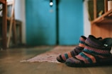 A pair of colorful knitted slippers with a striped design lies on a wooden floor. The background features a blurred interior setting with a turquoise wall and wooden furniture. A woven rug extends from the slippers toward the back of the room.