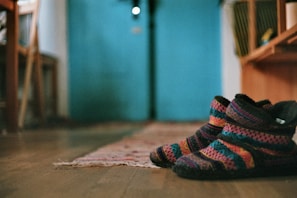 A pair of colorful knitted slippers with a striped design lies on a wooden floor. The background features a blurred interior setting with a turquoise wall and wooden furniture. A woven rug extends from the slippers toward the back of the room.