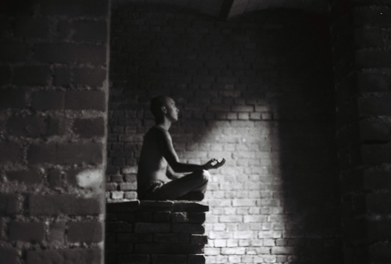 A person meditates in a dark, brick-walled space sitting cross-legged on a raised platform. Soft light illuminates their profile, creating a serene atmosphere.