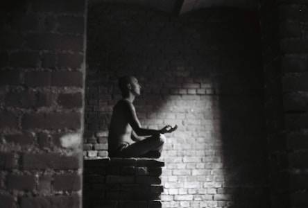 A person meditates in a dark, brick-walled space sitting cross-legged on a raised platform. Soft light illuminates their profile, creating a serene atmosphere.