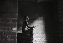 A person meditates in a dark, brick-walled space sitting cross-legged on a raised platform. Soft light illuminates their profile, creating a serene atmosphere.