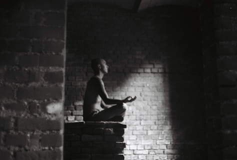 A person meditates in a dark, brick-walled space sitting cross-legged on a raised platform. Soft light illuminates their profile, creating a serene atmosphere.