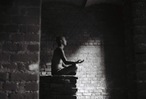 A person meditates in a dark, brick-walled space sitting cross-legged on a raised platform. Soft light illuminates their profile, creating a serene atmosphere.