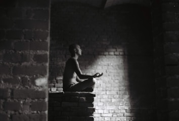 A person meditates in a dark, brick-walled space sitting cross-legged on a raised platform. Soft light illuminates their profile, creating a serene atmosphere.