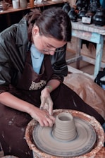 A person wearing glasses and a dark apron is working with clay on a pottery wheel. Their hands are shaping the clay as the wheel spins. In the background, there are various pottery items and tools on shelves.