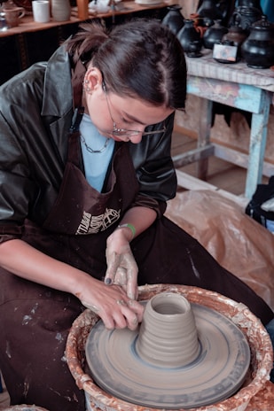 A person wearing glasses and a dark apron is working with clay on a pottery wheel. Their hands are shaping the clay as the wheel spins. In the background, there are various pottery items and tools on shelves.
