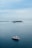 A large white and red yacht floats peacefully on a vast expanse of calm blue ocean. In the distance, a small lush island with several structures and palm trees is visible along with a long breakwater stretching across the horizon. The sky is overcast with soft, light gray clouds.