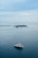 A large white and red yacht floats peacefully on a vast expanse of calm blue ocean. In the distance, a small lush island with several structures and palm trees is visible along with a long breakwater stretching across the horizon. The sky is overcast with soft, light gray clouds.