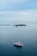 A large white and red yacht floats peacefully on a vast expanse of calm blue ocean. In the distance, a small lush island with several structures and palm trees is visible along with a long breakwater stretching across the horizon. The sky is overcast with soft, light gray clouds.