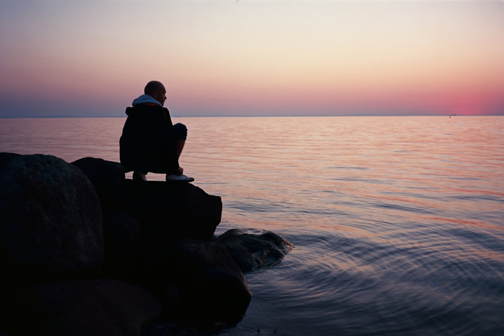 silhouette d’homme assis sur un rocher au milieu de la mer pendant le coucher du soleil