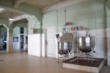 A large industrial kitchen with two massive stainless steel mixing bowls or vats. The room features pale green walls with white tile covering the lower half and a ceiling crisscrossed with exposed beams. Several canned goods and containers are neatly arranged on a shelf above the mixers. The space is well-lit with a lamp hanging from the ceiling.