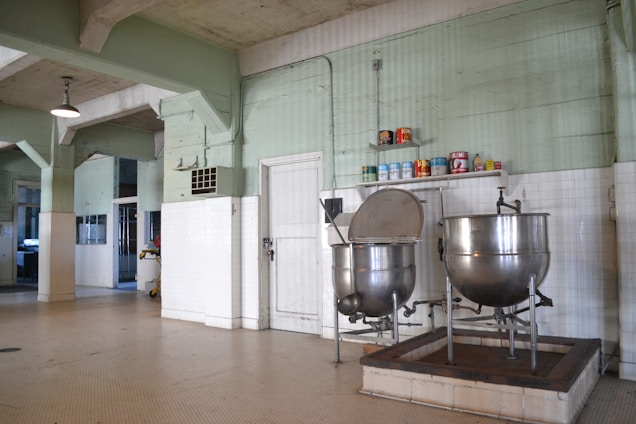 A large industrial kitchen with two massive stainless steel mixing bowls or vats. The room features pale green walls with white tile covering the lower half and a ceiling crisscrossed with exposed beams. Several canned goods and containers are neatly arranged on a shelf above the mixers. The space is well-lit with a lamp hanging from the ceiling.