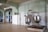 A large industrial kitchen with two massive stainless steel mixing bowls or vats. The room features pale green walls with white tile covering the lower half and a ceiling crisscrossed with exposed beams. Several canned goods and containers are neatly arranged on a shelf above the mixers. The space is well-lit with a lamp hanging from the ceiling.