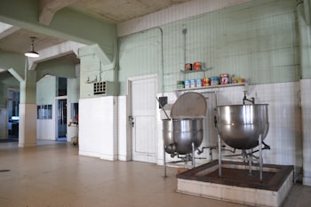 A large industrial kitchen with two massive stainless steel mixing bowls or vats. The room features pale green walls with white tile covering the lower half and a ceiling crisscrossed with exposed beams. Several canned goods and containers are neatly arranged on a shelf above the mixers. The space is well-lit with a lamp hanging from the ceiling.